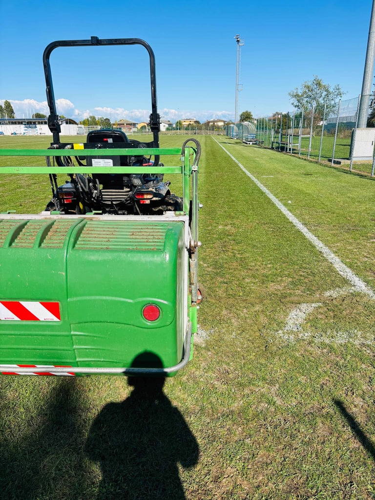 Green sports field maintenance machine on a soccer pitch with white line markings and stadium in background