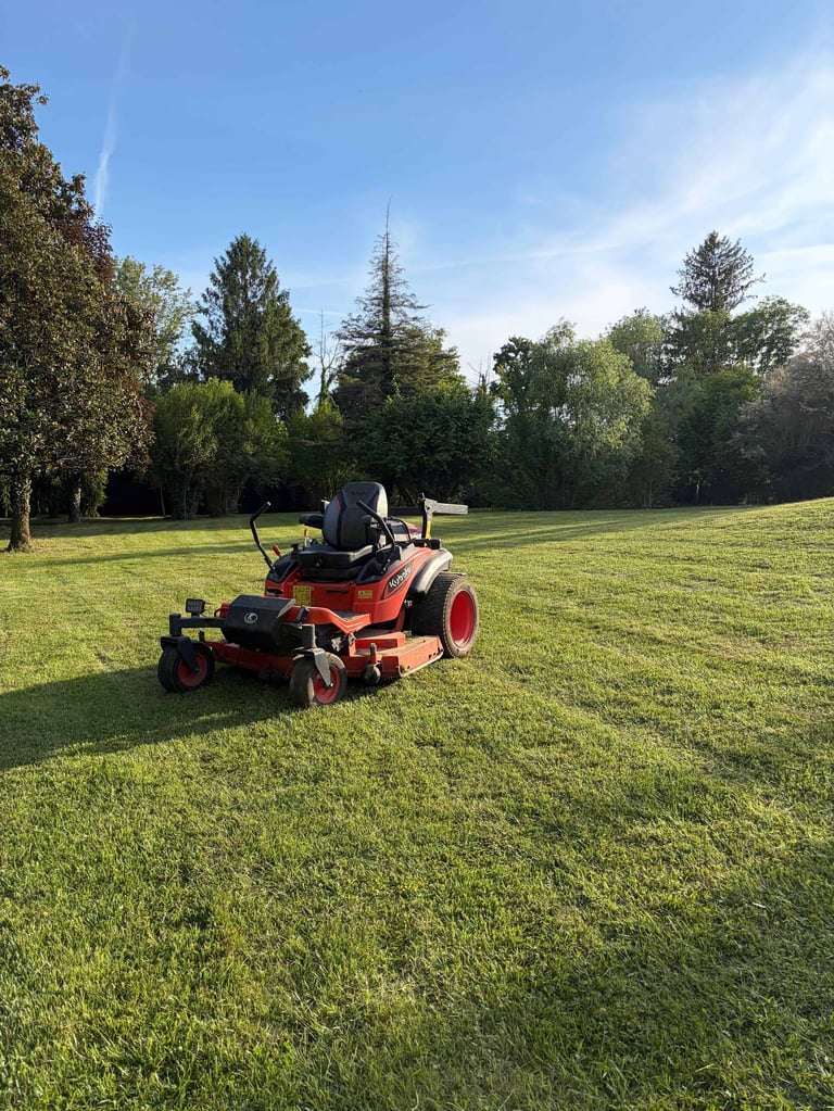 Red and black riding lawn mower on a grassy field with trees in the background under a clear blue sky